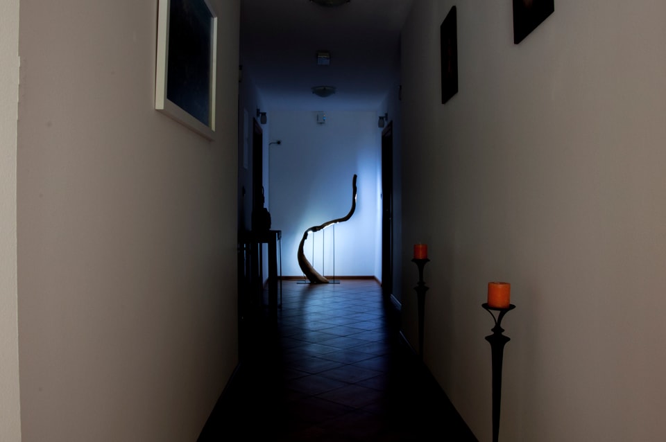 A narrow hallway is shown, featuring a striking light source illuminating a curved object against the wall. Candlesticks with orange candles line the floor, creating a soft glow. The floor is tiled, and artwork is displayed on the walls, contributing to the ambiance.
