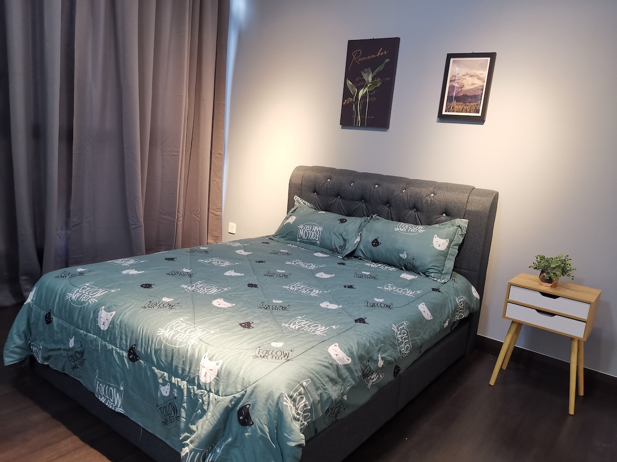 A spacious bed is dressed in a teal cover featuring playful graphics. Two decorative pillows rest at the head of the bed against a tufted gray headboard. Soft lighting highlights the room's neutral wall color, complemented by a small bedside table with a potted plant.