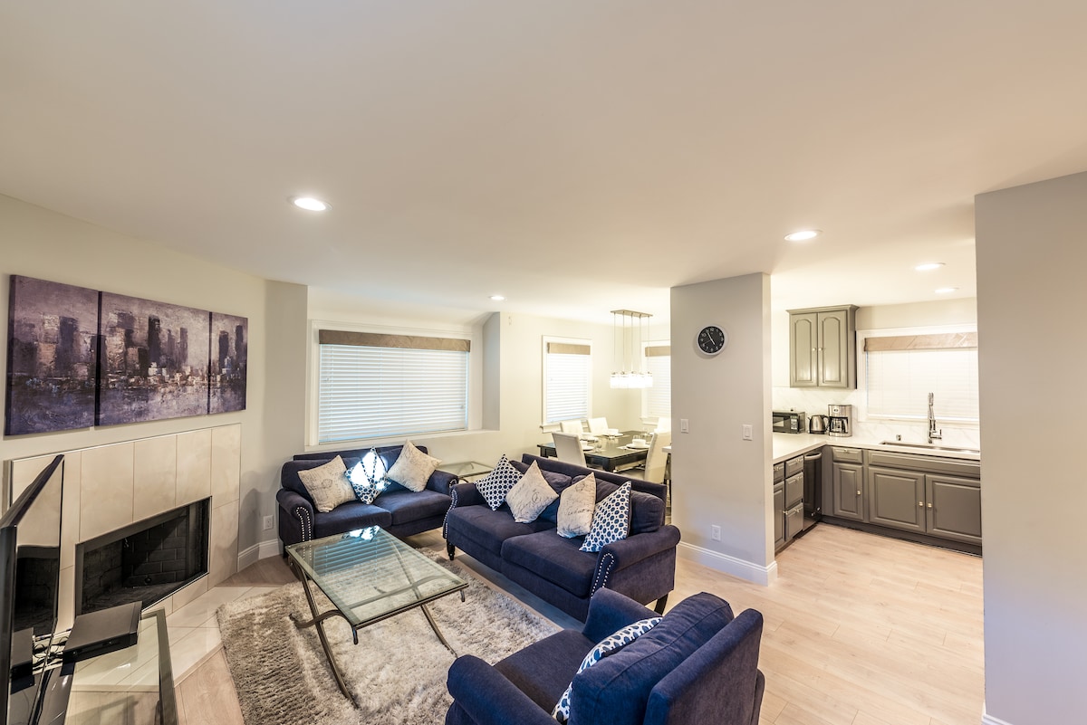 A spacious living area features a comfortable seating arrangement with dark blue sofas adorned with patterned cushions. A glass coffee table sits at the center. Light streams in through multiple windows, highlighting the modern kitchen area with stainless steel appliances and elegant cabinetry.