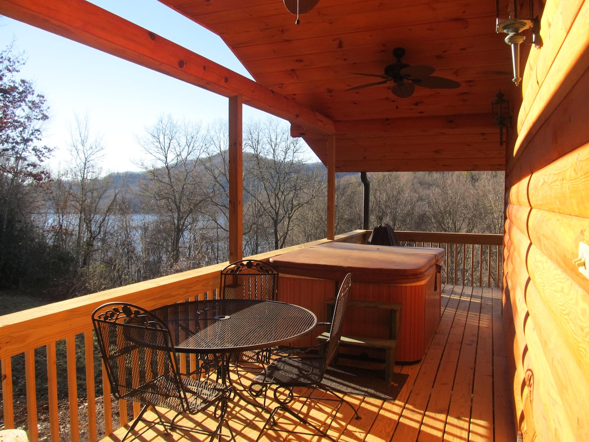 A wooden deck features a hot tub and a dining area with a round table and four chairs. The view of the lake and surrounding trees can be seen in the background, illuminated by natural light.