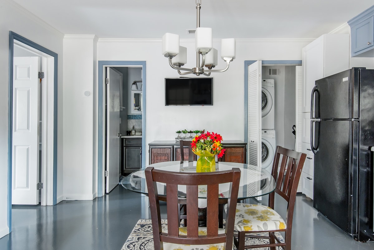 A dining area features a round glass table surrounded by four wooden chairs, with a vase of colorful flowers at its center. A wall-mounted television is visible above a stylish cabinet, while a doorway leads to a laundry area opposite the kitchen entrance.