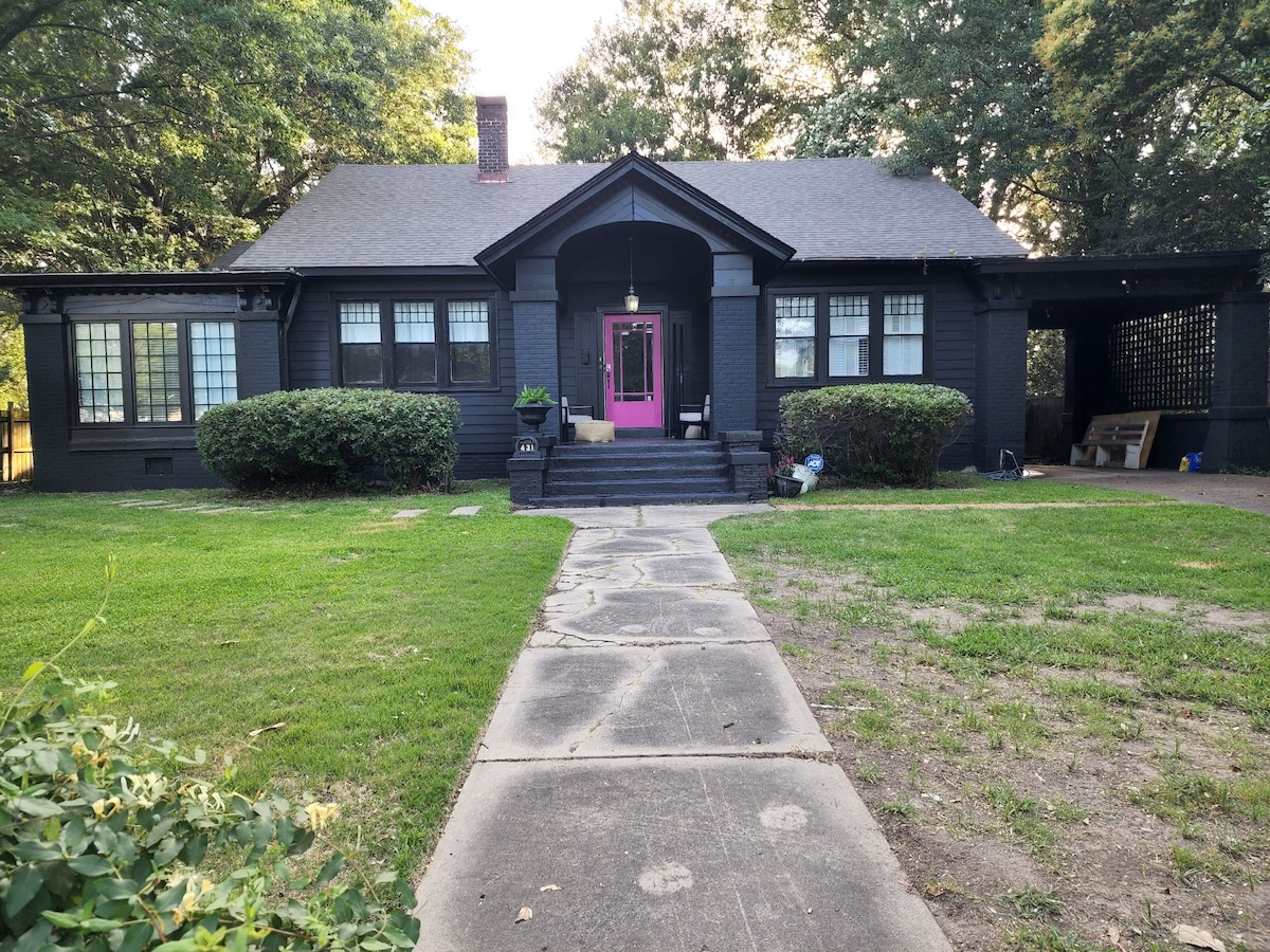 The exterior of a historic black house is visible, featuring a distinctive pink door at the center. Bay windows on either side are set within a manicured landscape. A concrete pathway leads to the entrance, bordered by neatly trimmed shrubs and a grassy yard.