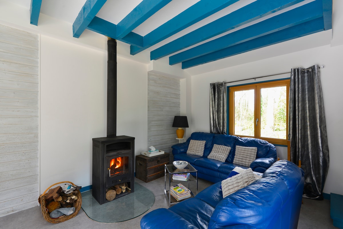 A cozy living room features a modern wood stove set on a circular platform. Two blue sofas are arranged around a glass coffee table, showcasing magazines and a small decorative item. Natural light enters through a window adorned with gray curtains.