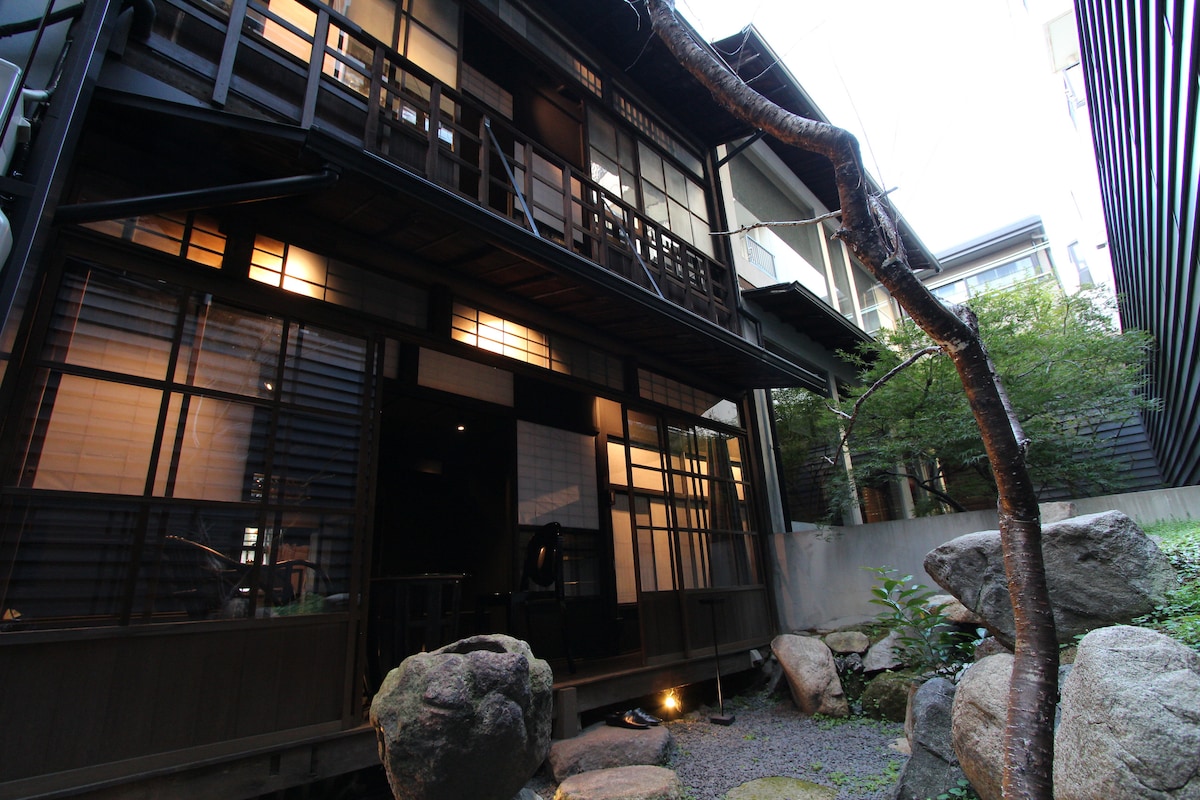 An elegantly renovated traditional Japanese house is showcased, featuring a well-maintained garden with smooth stones and greenery. The structure is characterized by wooden beams and large glass windows, allowing natural light to enter while preserving the classic aesthetic.