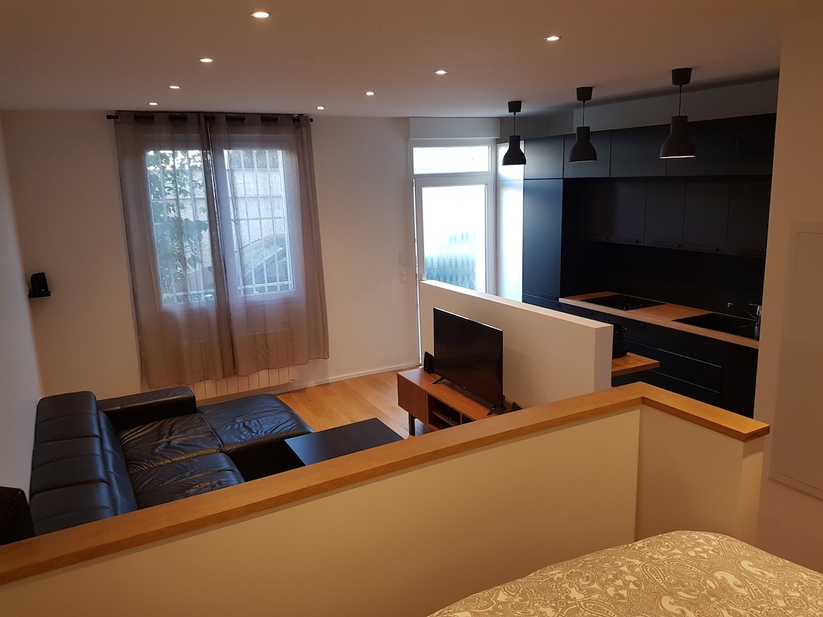 A spacious living area is presented, featuring a black sectional sofa and a wooden coffee table. A flat-screen television sits on a console, with ambient light provided by ceiling lights. The modern kitchen, equipped with sleek cabinetry and wooden countertops, is visible in the background. Natural light streams in through expansive windows framed with light curtains.