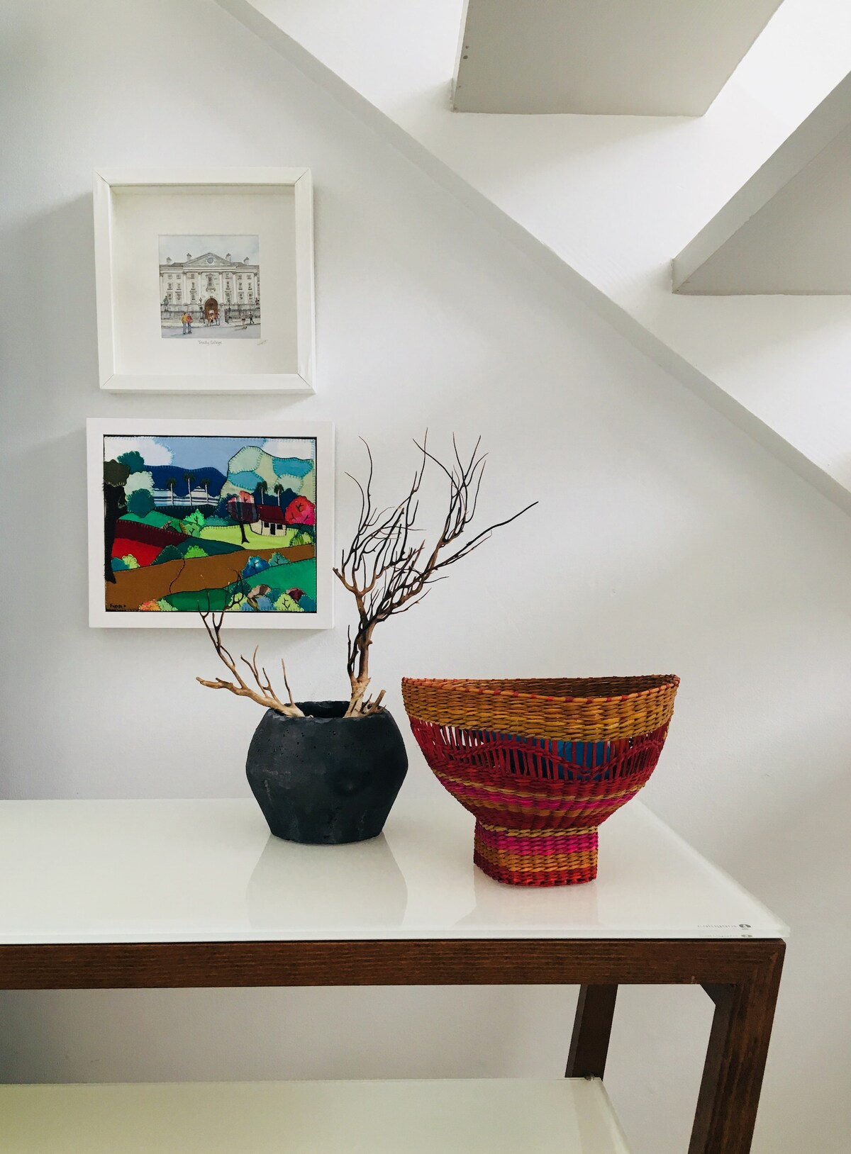 A modern side table is positioned against a white wall, featuring two decorative pieces. A black ceramic vase with branches stands beside a colorful woven basket. Above, framed art pieces in vibrant colors add visual interest to the space.