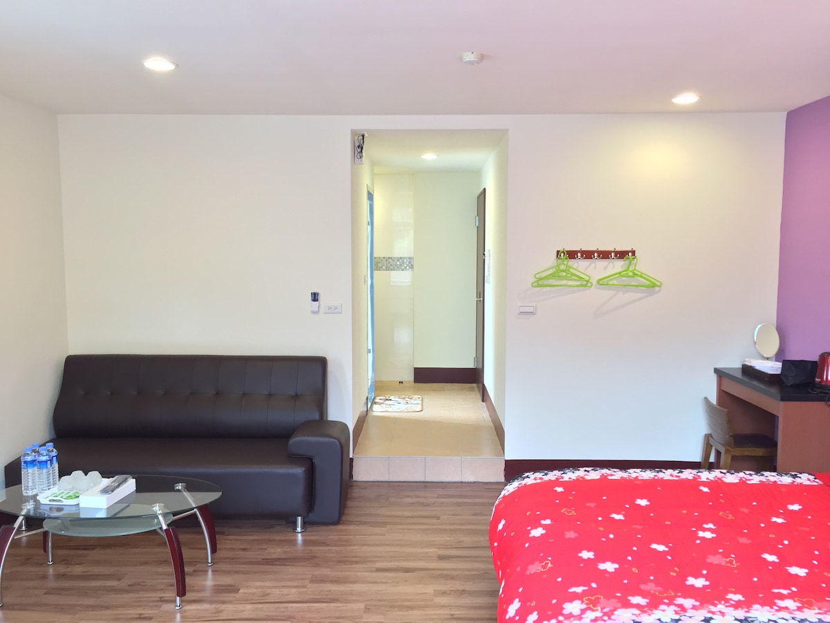 A tidy, well-lit room features dark wood flooring and a vibrant purple accent wall. A spacious bed is adorned with a red floral blanket. A seating area includes a black sofa and a glass coffee table. A doorway leads to a bathroom, visible in the background.