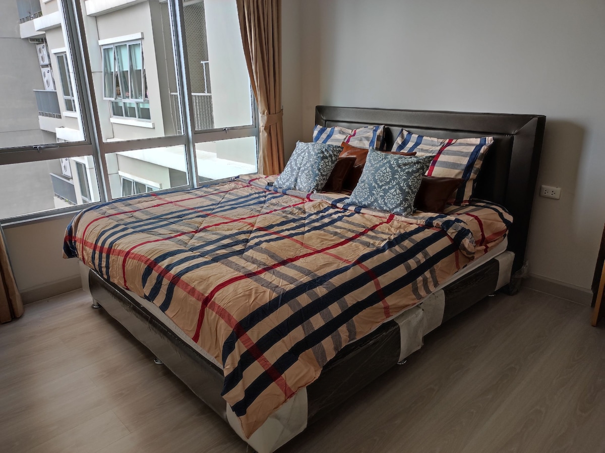 A spacious sleeping area features a large bed with a patterned comforter and an assortment of decorative pillows. Sunlight streams through large windows, providing natural light to the room, which is complemented by neutral-toned walls and wooden flooring.