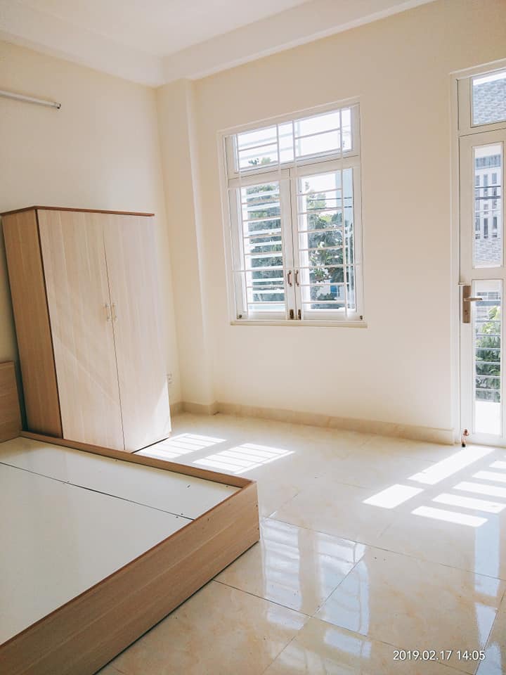 A bright room features large windows allowing ample natural light to fill the space. A simple wardrobe stands against the wall, while a bed frame is positioned adjacent to it. The light-colored tiles enhance the airy feel of the interior.