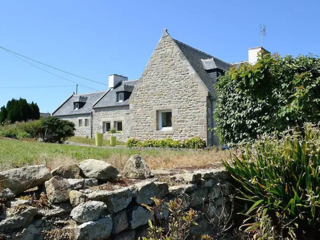 A traditional Breton stone house is depicted, featuring a charming gabled roof and decorative window trim. Surrounding greenery and flowering plants enhance the exterior, with a well-maintained garden area visible. The property's rustic stone wall adds character to the landscape.