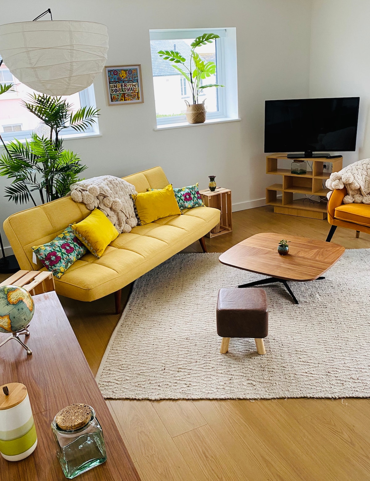 A bright living area is showcased, featuring a yellow sofa adorned with colorful pillows. A wooden coffee table anchors the space, accompanied by a small brown stool. Natural light fills the room through two windows, complemented by indoor plants and minimalistic decor.