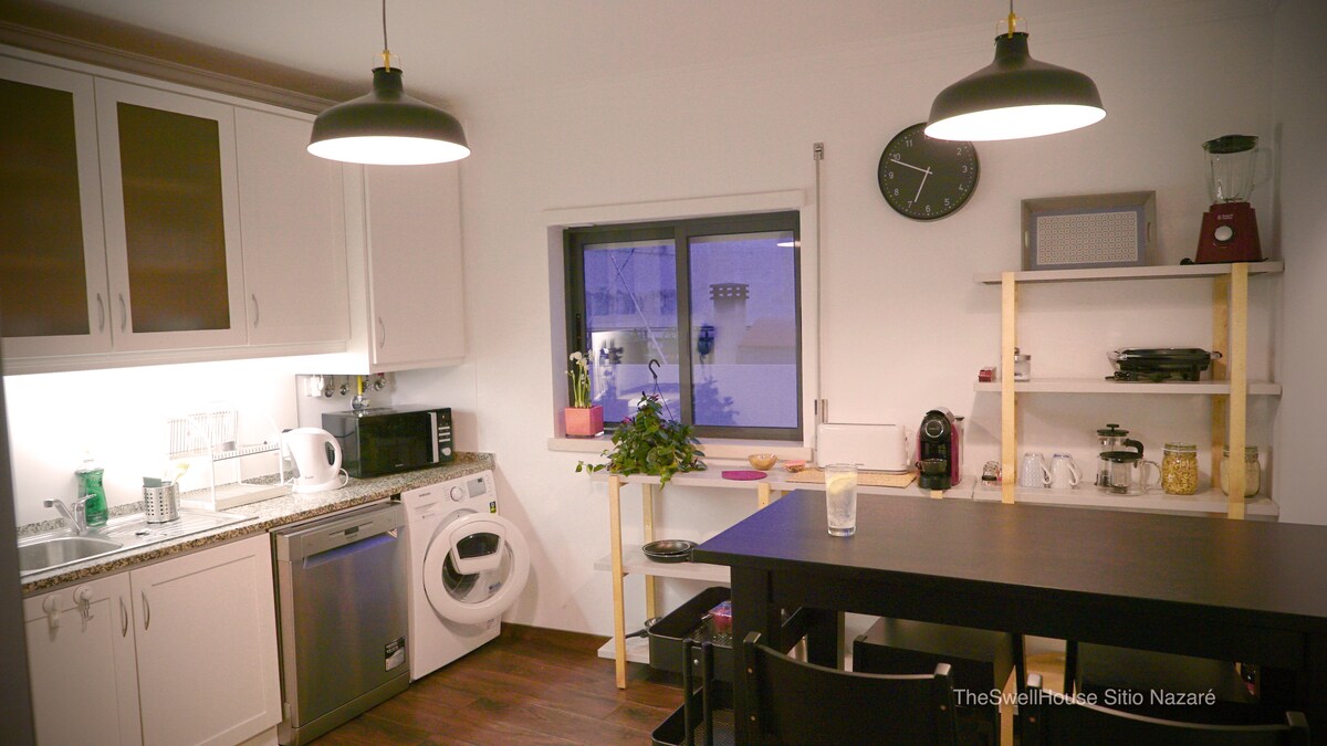 The kitchen area features modern cabinetry, a granite countertop, and essential appliances, including a washing machine and microwave. A dining table for four is positioned nearby, with minimalistic shelving displaying kitchenware. Natural light filters in through a window, adding warmth to the space.