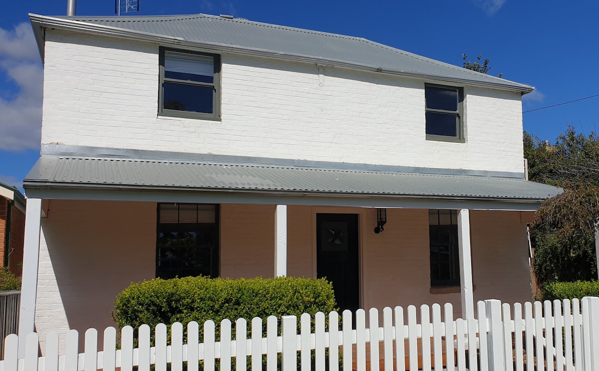 The cottage features a classic two-story facade, highlighted by a light-colored paint scheme. A metal roof and front porch with columns are present. A white picket fence surrounds a neat green hedge, complementing the structured appearance of the home.