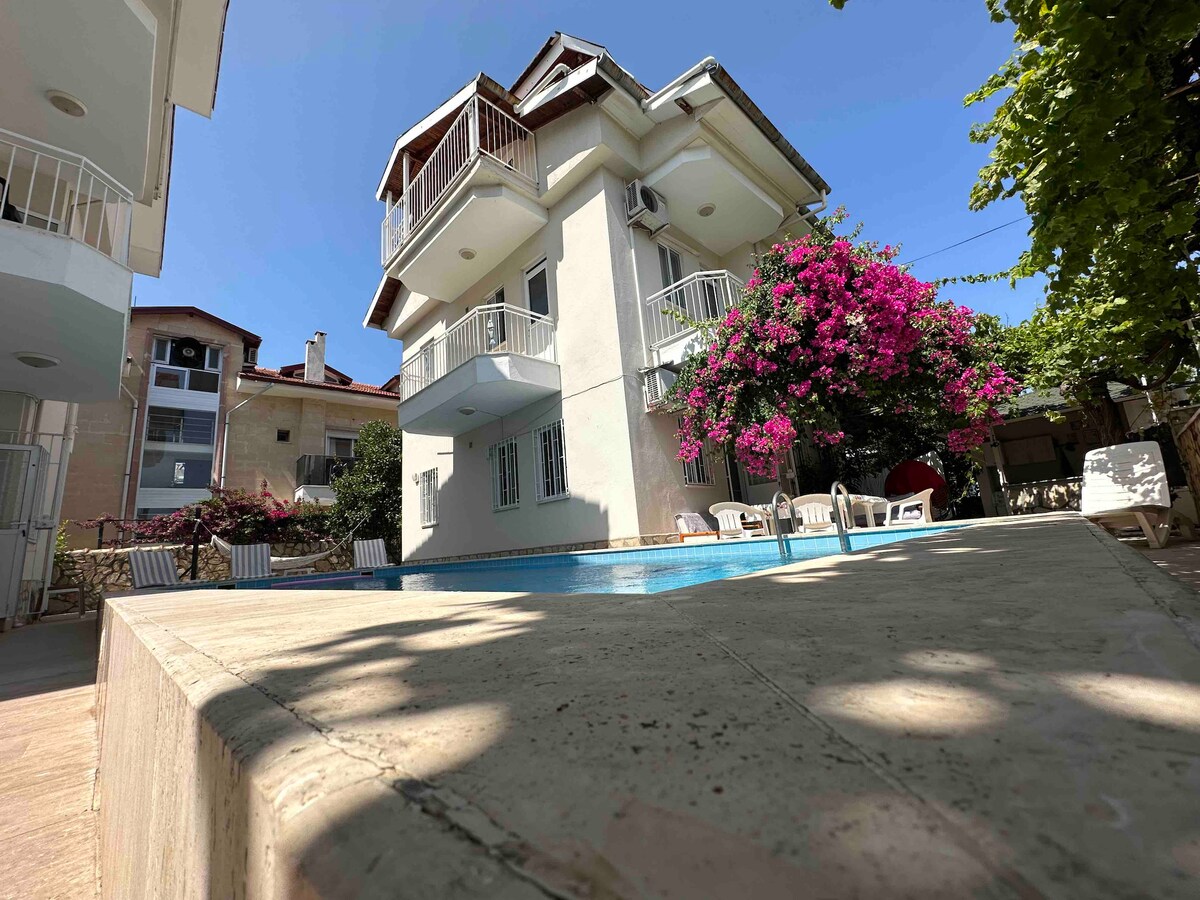 The exterior view features a three-story building with balconies overlooking a pool area. Lush pink flowering plants adorn the surroundings, while sun loungers are positioned near the pool. Bright sunlight illuminates the scene, creating a relaxing outdoor space.
