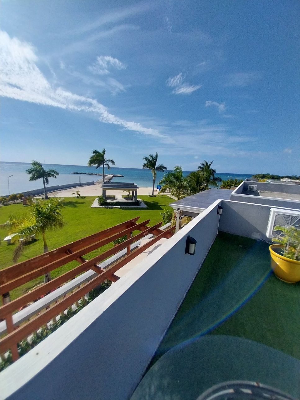 A balcony view reveals a spacious patio area with palm trees and a manicured lawn leading to a sandy beach. The ocean glimmers under a clear blue sky, offering a serene coastal atmosphere.
