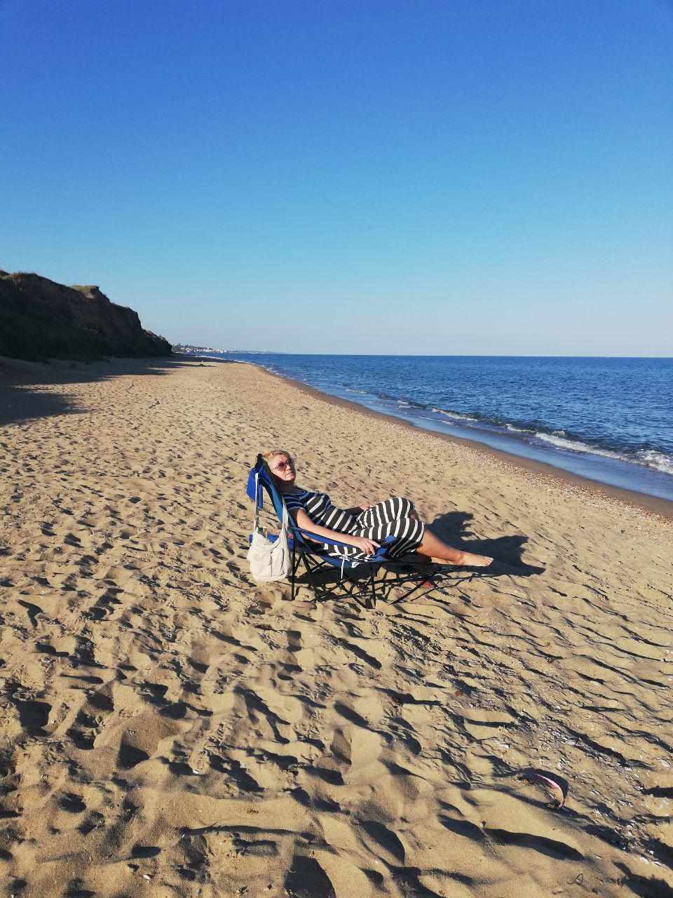 A sandy beach is shown under a clear blue sky, with gentle waves lapping at the shore. A folding chair is positioned near the water's edge, suggesting a serene space for relaxation. The scene conveys tranquility and the natural beauty of the coastline.