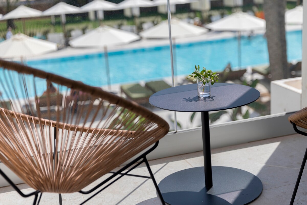 A relaxed seating area is highlighted by a modern chair and a small black table featuring a slender vase with greenery. The background reveals a pool area adorned with umbrellas, suggesting a refreshing atmosphere for guests.