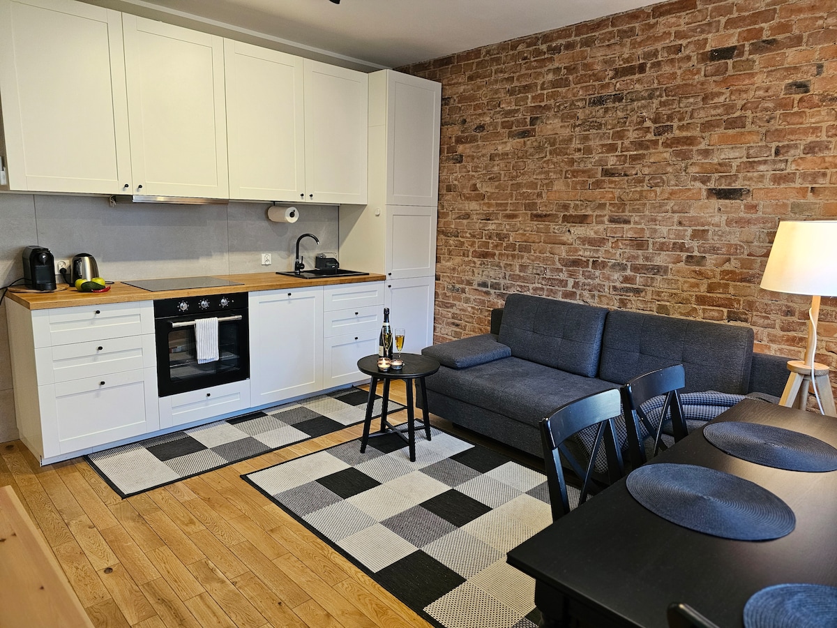 The kitchen area features modern white cabinetry paired with a wooden countertop. A comfortable gray sofa is positioned in front of a small black table. A dining table with black chairs occupies one corner, while textured brick walls add warmth to the space.