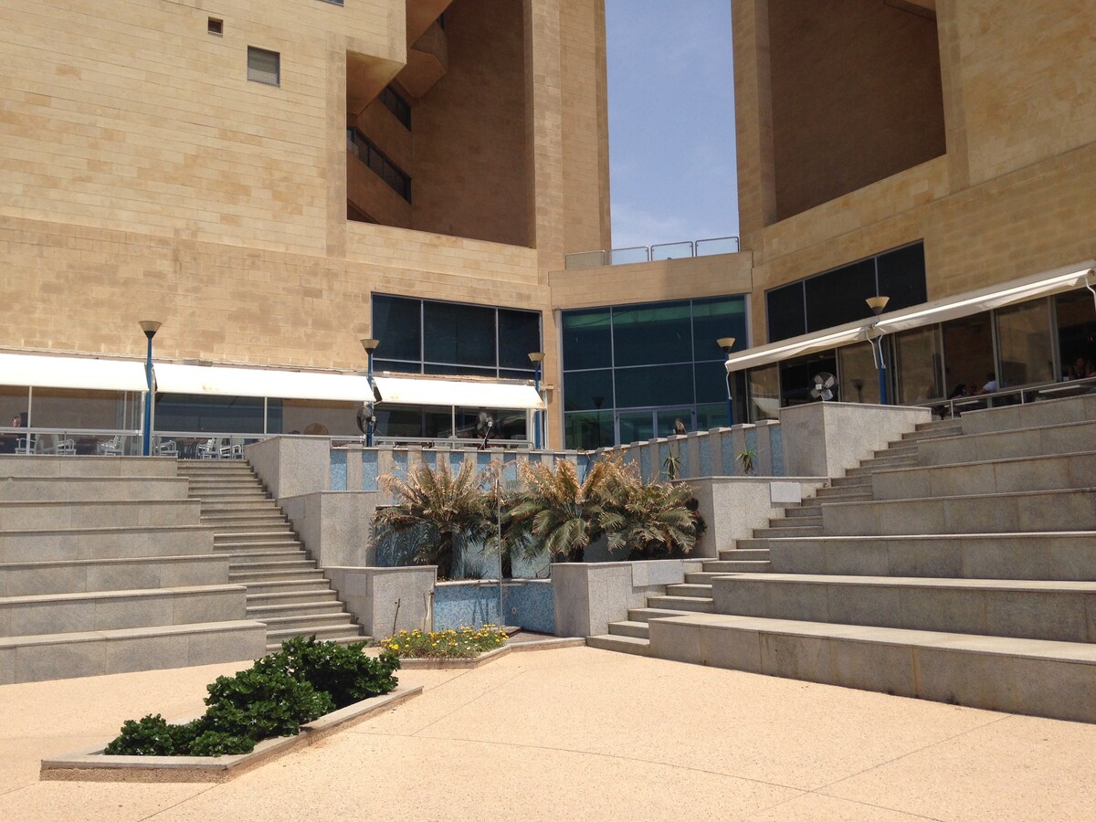 The exterior area is highlighted by a series of tiered steps leading down to a small water feature surrounded by lush greenery. Large windows are visible in the background, reflecting sunlight. Several outdoor seating areas are positioned along the terrace.