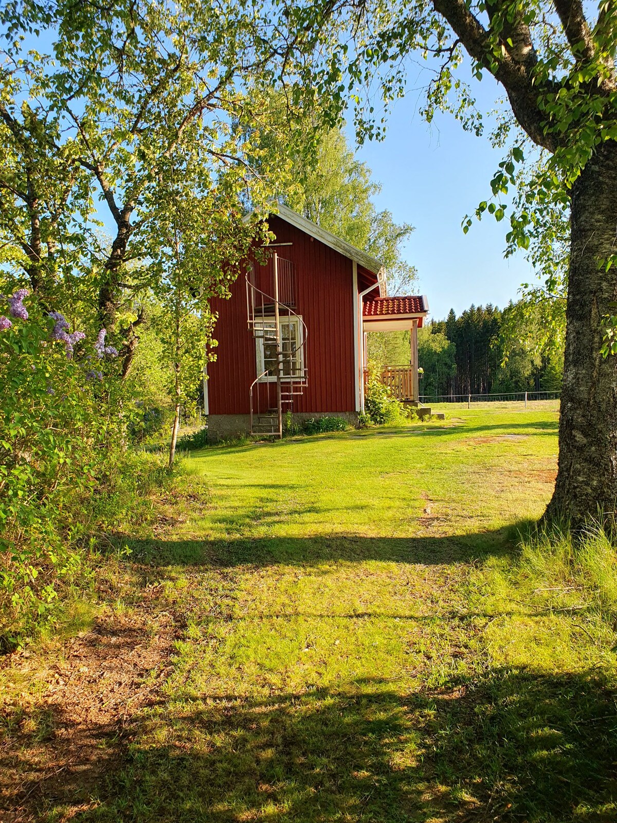 A charming red cabin is positioned in a tranquil outdoor setting, surrounded by trees and greenery. The structure features a spiral staircase leading to a balcony, offering a blend of natural beauty and rustic architecture. Sunlight filters through the leaves, adding warmth to the scene.