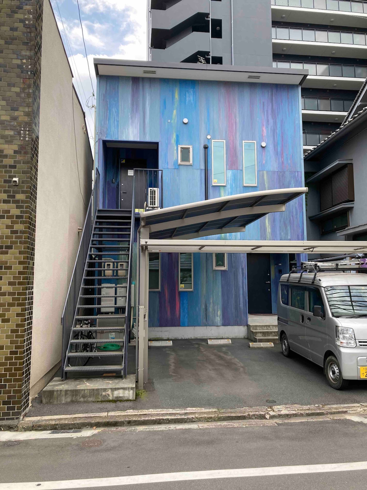 A modern two-story building features a striking exterior in shades of blue and purple, complemented by three vertical windows. External stairs lead to the second floor, with a carport providing shelter for a vehicle. Surrounding structures and a paved pathway complete the urban setting.