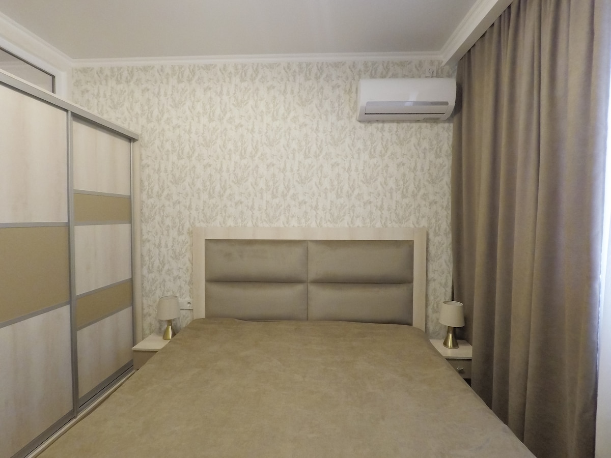 A spacious bed with a soft, neutral-colored headboard is centered against a floral-patterned wall. Bedside tables with lamps are positioned on either side, while a large wardrobe is visible to the left. Soft curtains frame a window, contributing to a calm ambiance.