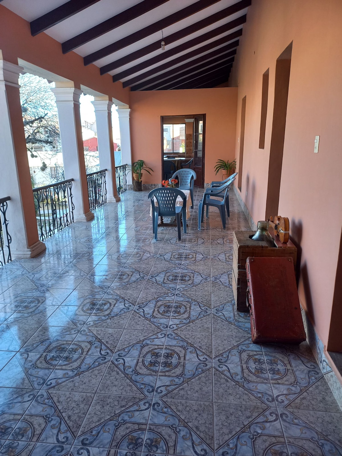 A spacious terrace is lined with decorative columns, featuring tiled flooring. Two simple chairs are arranged around a small table, with a potted plant visible nearby. Natural light illuminates the area, enhancing the warm tones of the surrounding walls.