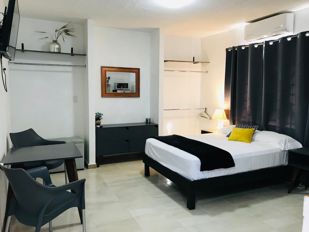 A well-lit room features a spacious bed with neatly arranged white linens and a decorative black throw. A flat-screen TV is mounted on a wall. Gray chairs and a modern table complement a dresser opposite the bed, while a decorative plant adds a touch of warmth.