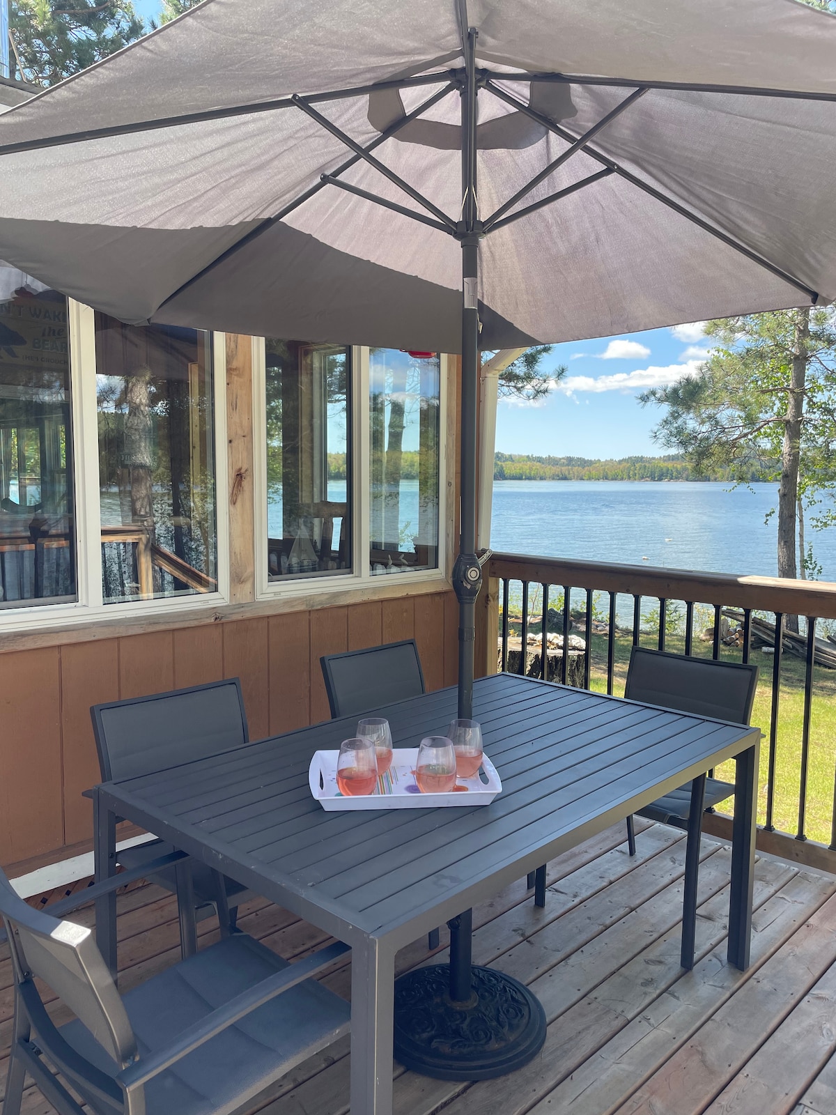 An outdoor dining area is visible, featuring a gray table surrounded by five chairs under a large umbrella. A scenic view of the lake is framed by trees, offering a serene spot for enjoying meals or beverages.