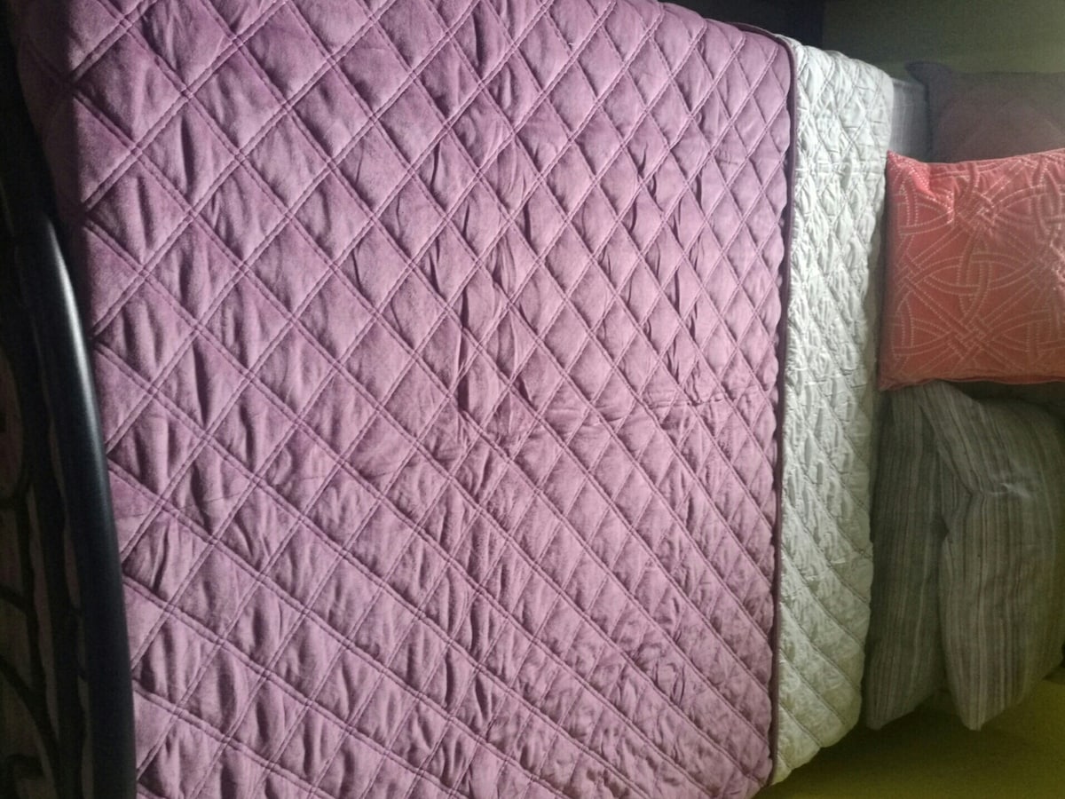 A spacious bed is covered with a light purple quilted comforter. Several decorative pillows in varying patterns and colors are arranged at the head of the bed, complemented by neutral-tone bed linens. The soft ambiance of the sleeping area is enhanced by natural light.