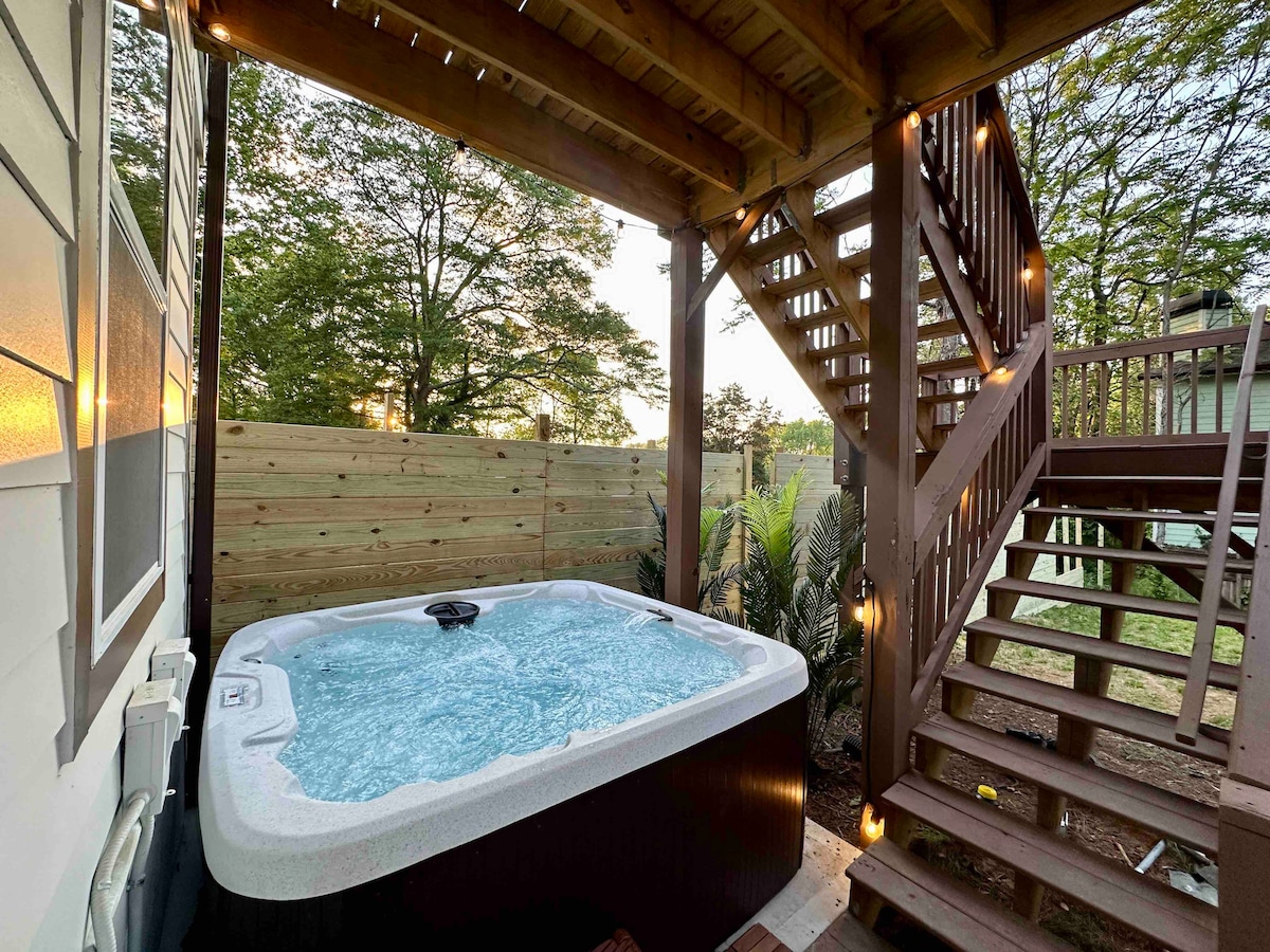 A private hot tub is nestled under a wooden deck, surrounded by lush greenery and modern wooden fencing. Soft lighting accents the space, creating a serene ambiance. Stairs lead up from the deck, providing access to the upper level of the property.