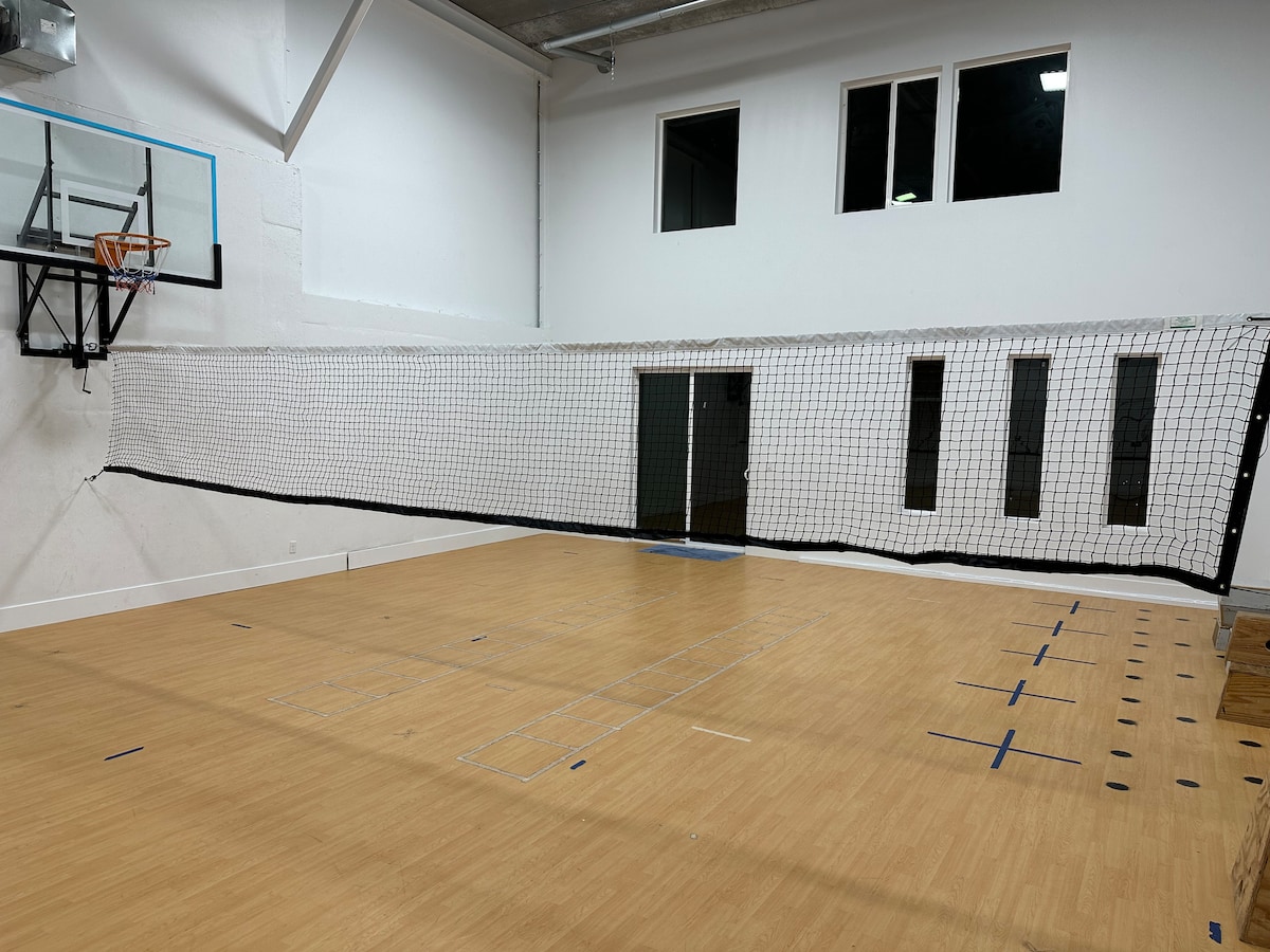 An indoor sports court is displayed, featuring a basketball hoop and a volleyball net. The wooden flooring is marked for various sports activities. Bright lights overhead illuminate the space, while large windows provide natural light, contributing to an open and airy atmosphere.