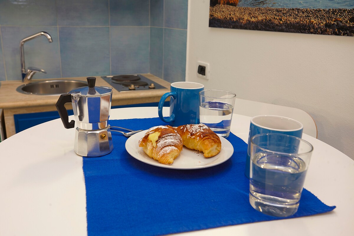 A round dining table is set with a blue tablecloth. Two glasses of water and two blue mugs are placed beside a plate of pastries, which are lightly dusted with powdered sugar. A stainless steel coffee maker is positioned nearby, while the background features kitchenette amenities.
