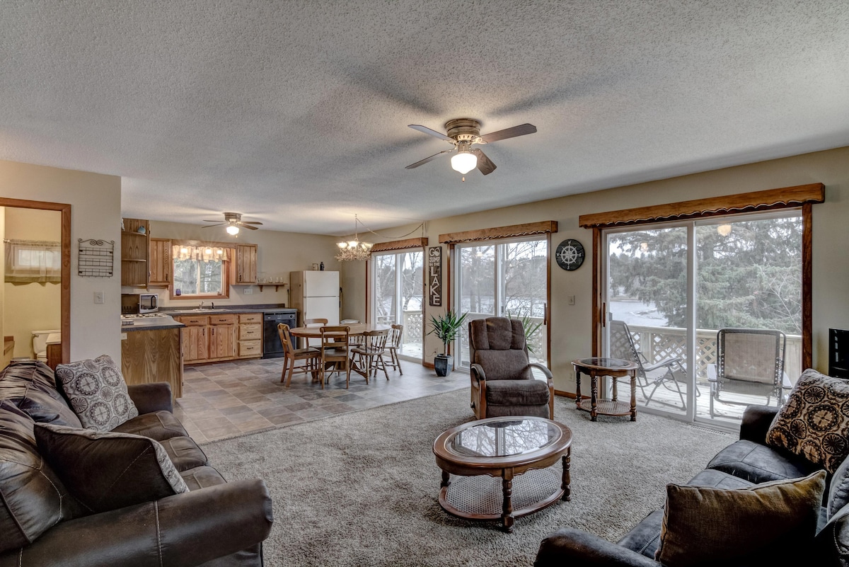 A spacious living area is furnished with comfortable seating, including a dark leather sofa and armchairs. Natural light fills the room through large windows, which offer views of the outdoors. A dining area is visible in an adjacent space, equipped with a table and chairs.