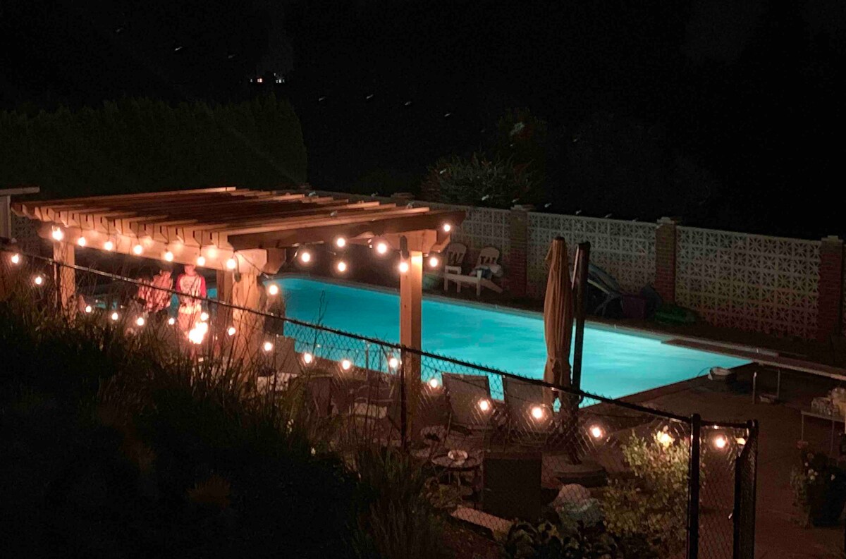 The nighttime scene depicts a tranquil pool area illuminated by soft string lights. A wooden pergola frames the space, providing a cozy gathering spot. The pool glows invitingly turquoise, surrounded by lounge chairs and a fence, creating a private and serene outdoor escape.
