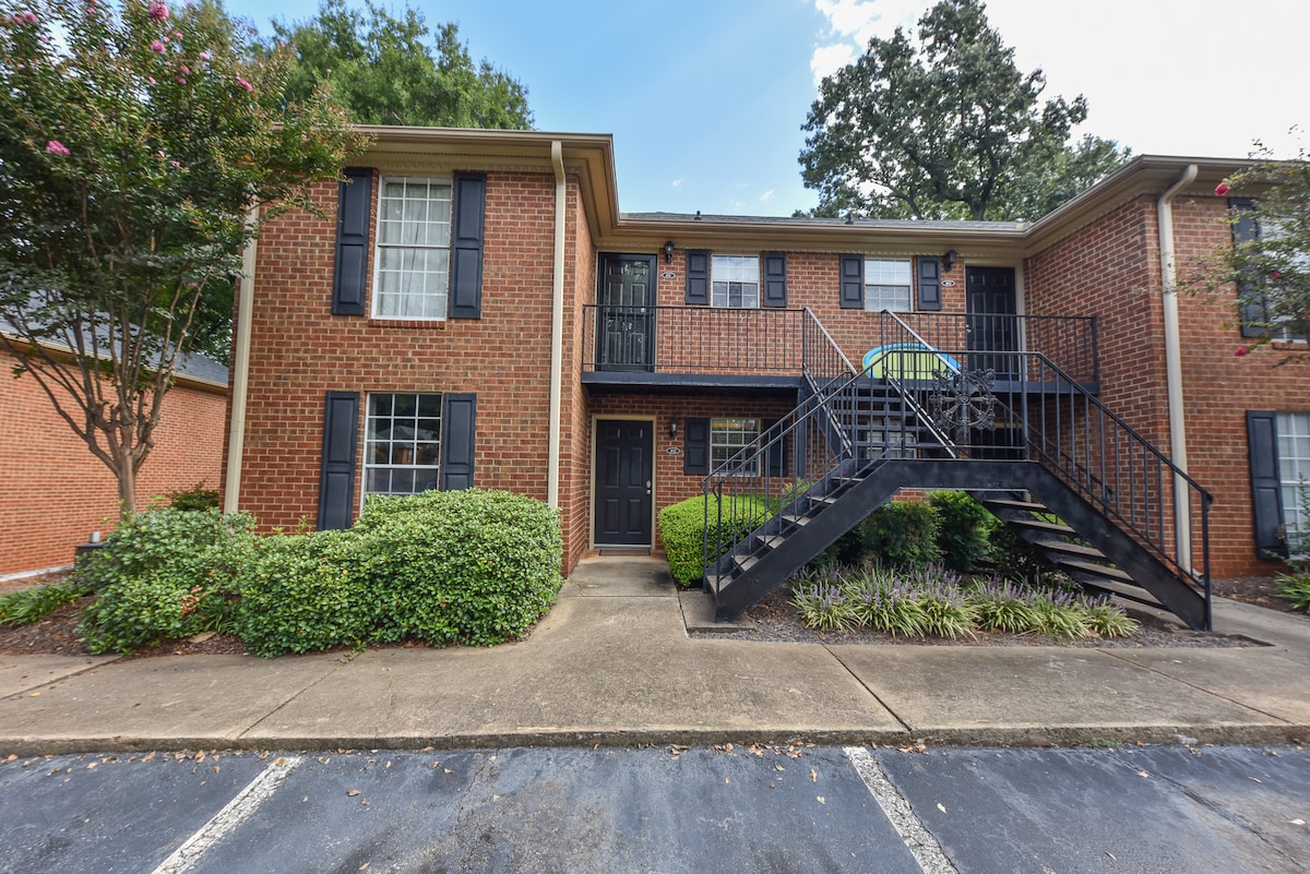 The exterior of a two-story brick condo is presented, featuring a mix of green shrubbery and concrete pathways. A staircase leads to a balcony entrance, which is adorned with a small seating area. Windows with dark shutters are framed by a tree with pink blooms.