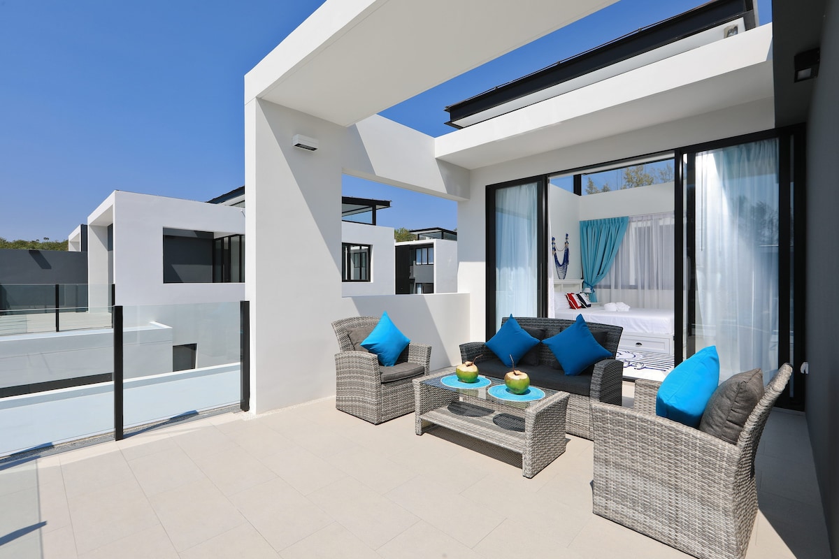 An outdoor terrace offers a seating arrangement featuring woven chairs and a coffee table adorned with decorative items. Bright blue cushions add a pop of color against the neutral tones. The view includes a glimpse of the interior's inviting sleeping area through large glass doors.