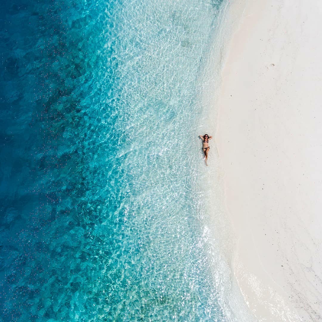 A tranquil beach scene captures soft white sand meeting crystal-clear turquoise waters. Gentle waves lap against the shore, creating a serene atmosphere. In the water, a person can be seen relaxing, enjoying the peaceful coastal environment.