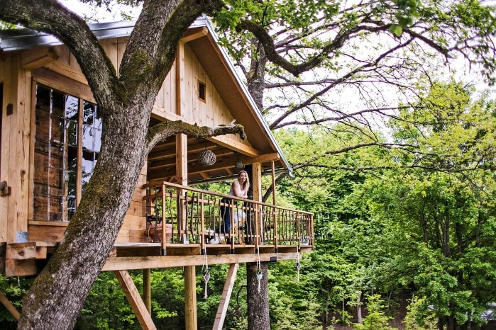 A wooden treehouse is perched among lush green trees, featuring a spacious balcony with wooden railings. A person is seen standing on the balcony, surrounded by nature. The cabin showcases rustic architecture with large windows, blending harmoniously into the serene environment.