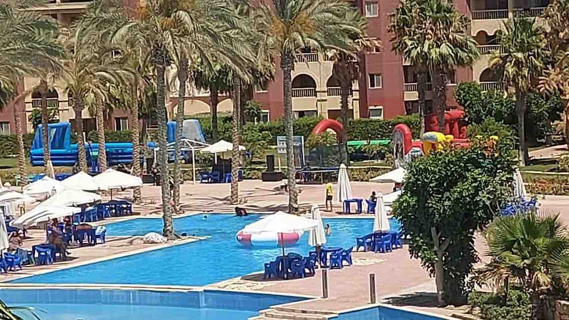 A large pool area is surrounded by palm trees and features numerous umbrellas over blue lounge chairs. Children’s play equipment is visible in the background, and vibrant inflatable structures add color to the recreational space.