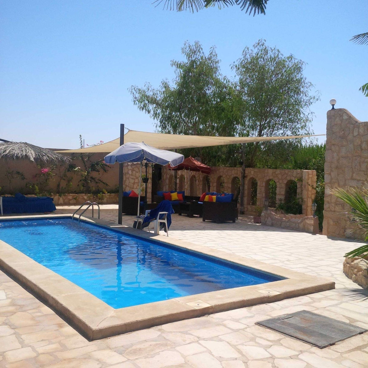 An inviting outdoor area features a swimming pool surrounded by a stone patio. Lounge chairs and a shaded seating area with colorful cushions provide spaces for relaxation. Lush greenery and decorative walls enhance the tranquil setting under clear blue skies.