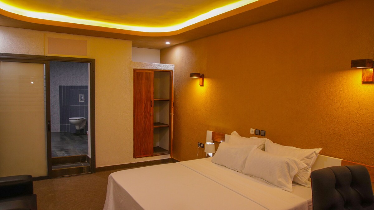 A spacious room features a comfortable bed with white bedding. Soft yellow lighting outlines the walls, creating a soothing ambiance. A glass door leads to a bathroom area, while a wooden closet provides storage. The neutral color palette enhances the room's welcoming atmosphere.