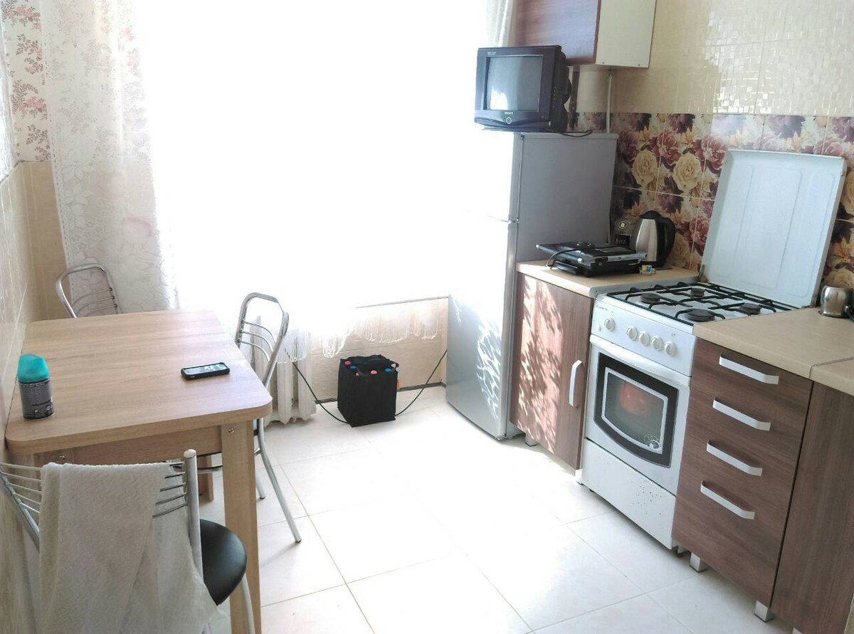 A kitchen area is illuminated by natural light from a large window. It features a modern stove, refrigerator, and microwave, along with a compact dining table and two chairs. A floral backsplash adds a touch of character to the otherwise neutral color scheme.