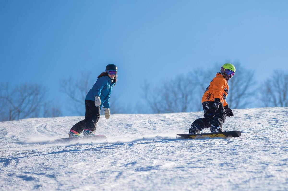 Two snowboarders navigate down a snowy slope. The person in a blue jacket follows closely behind another in an orange jacket. Bright blue skies and a backdrop of bare trees enhance the wintry scene.
