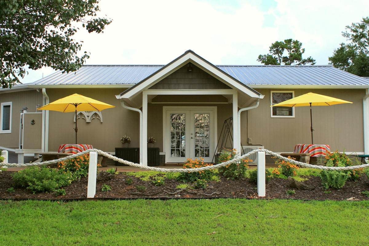 The exterior of the guest house is showcased with a charming entrance featuring double doors. Two yellow umbrellas provide shade over seating areas adorned with striped blankets. Colorful flowers and a decorative rope border enhance the inviting atmosphere of the landscaped yard.
