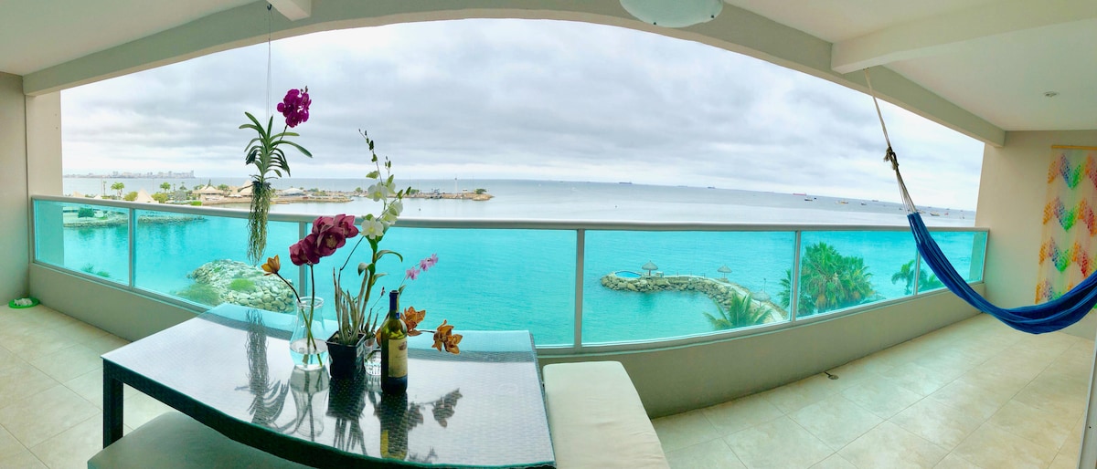 A spacious balcony is shown, featuring a glass table adorned with flowers and a view of the serene blue ocean. A vibrant blue hammock is visible, gracefully hanging beside the table, while the soft colors of the sky complement the tranquil waters.