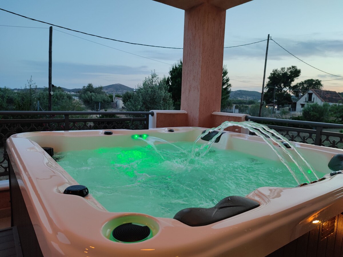 A spacious jacuzzi is positioned on the veranda, featuring illuminated jets and a waterfall. The water emits a calming green glow, surrounded by a scenic view of the landscape. The setting provides an inviting space for relaxation, enhanced by the evening sky.