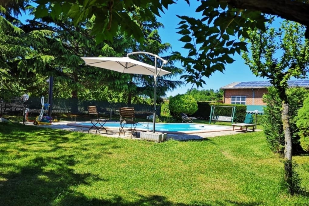 The image showcases a well-maintained outdoor area featuring a swimming pool surrounded by lush greenery. Lounge chairs are arranged on the pool deck, and a large umbrella provides shade. Nearby, a swing set is visible in a grassy space, enhancing the tranquil setting.