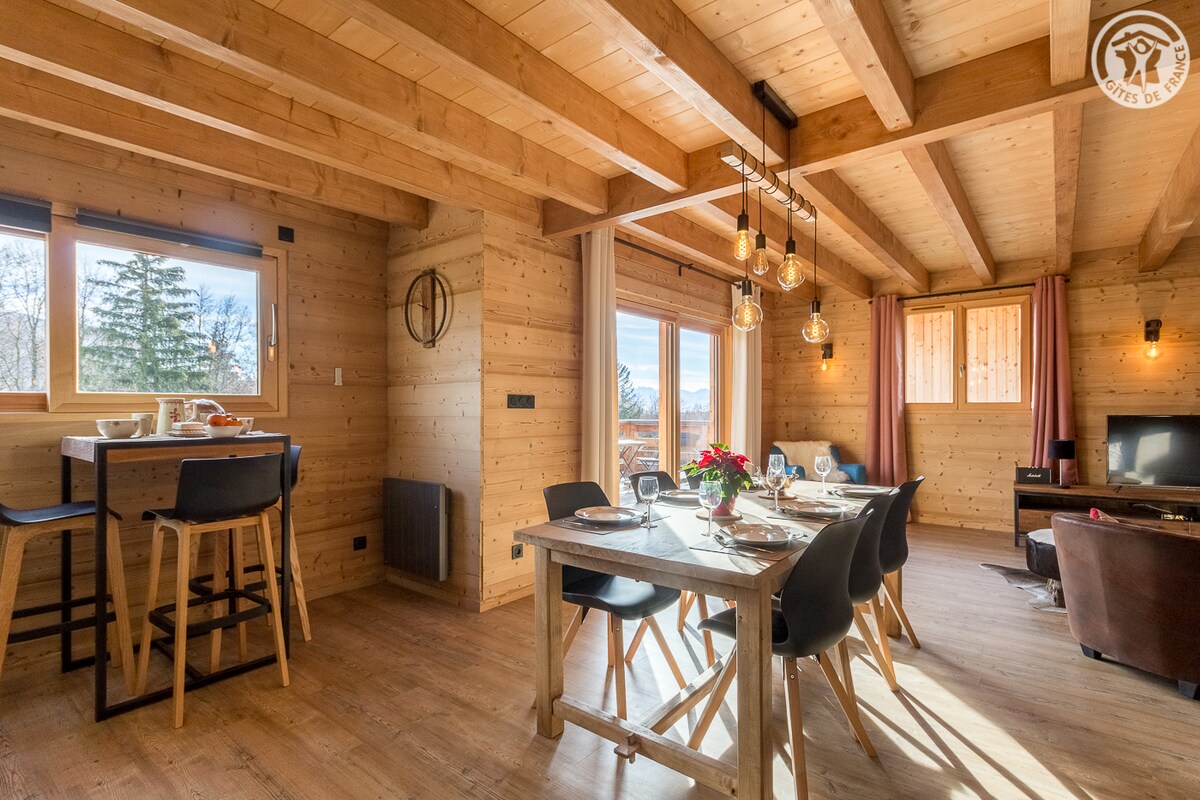 A cozy dining area features a wooden table surrounded by six chairs, set for a meal. Large windows offer natural light and views of the outdoors. Exposed wooden beams add warmth to the space, complemented by soft furnishings and modern lighting fixtures.
