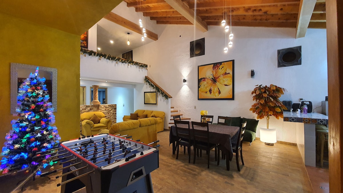 An open living space features a foosball table and a dining area with a large table. Colorful artwork adorns the walls, while a decorative tree adds holiday flair. Soft furnishings create a relaxed atmosphere, complemented by rustic wooden beams overhead.